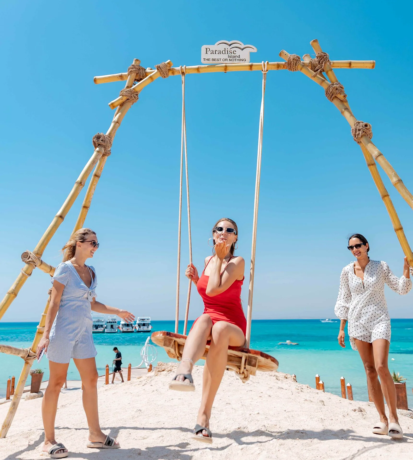A timber swing set on the sand at the shoreline, with guests beside it and the sea behind.