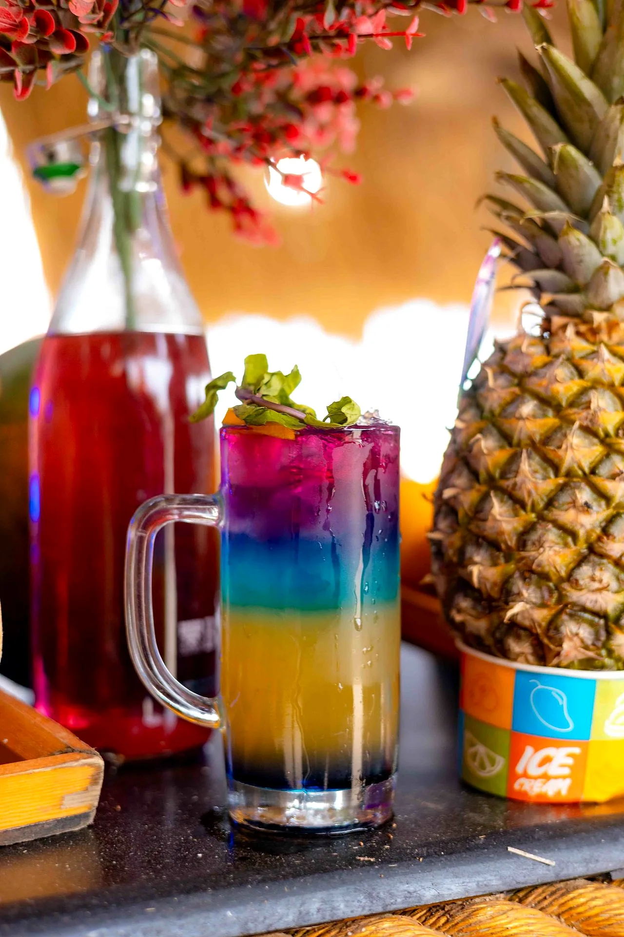 A tall layered cocktail and a jug of fruit punch on a bar top, with fresh pineapple behind.